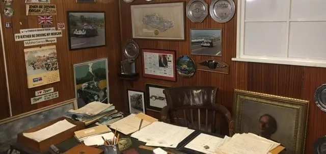 A wood-panelled office wall crowded with racing photographs, plates, stickers and a calendar, above a desk piled with papers and a leather chair.