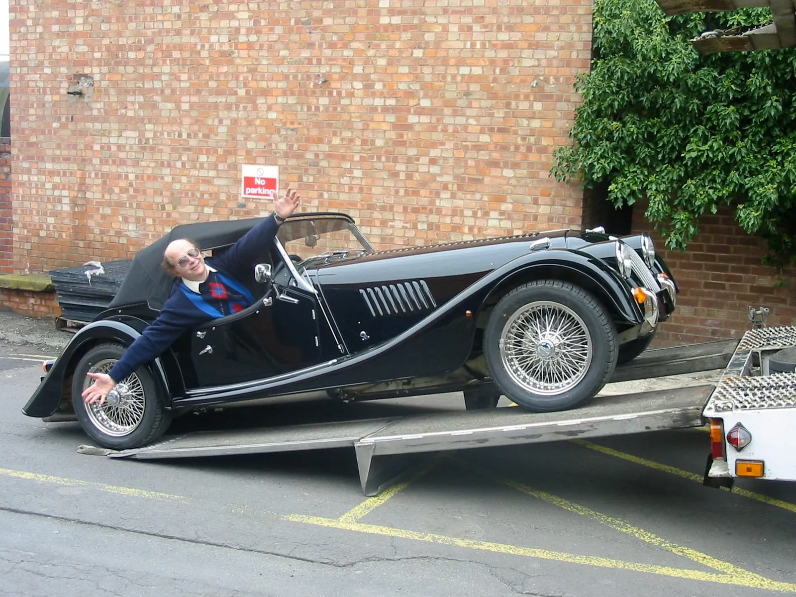 A man waves from the driver’s seat of a black Plus 8 as it is driven onto a trailer ramp, in front of a brick wall