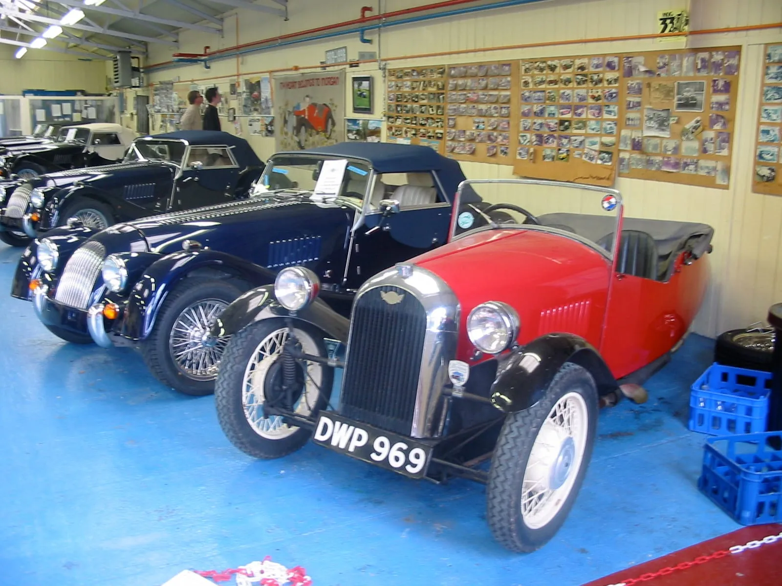 Old Morgans, including a red three-wheeler and two blue four-wheelers, parked in a workshop hung with photographs