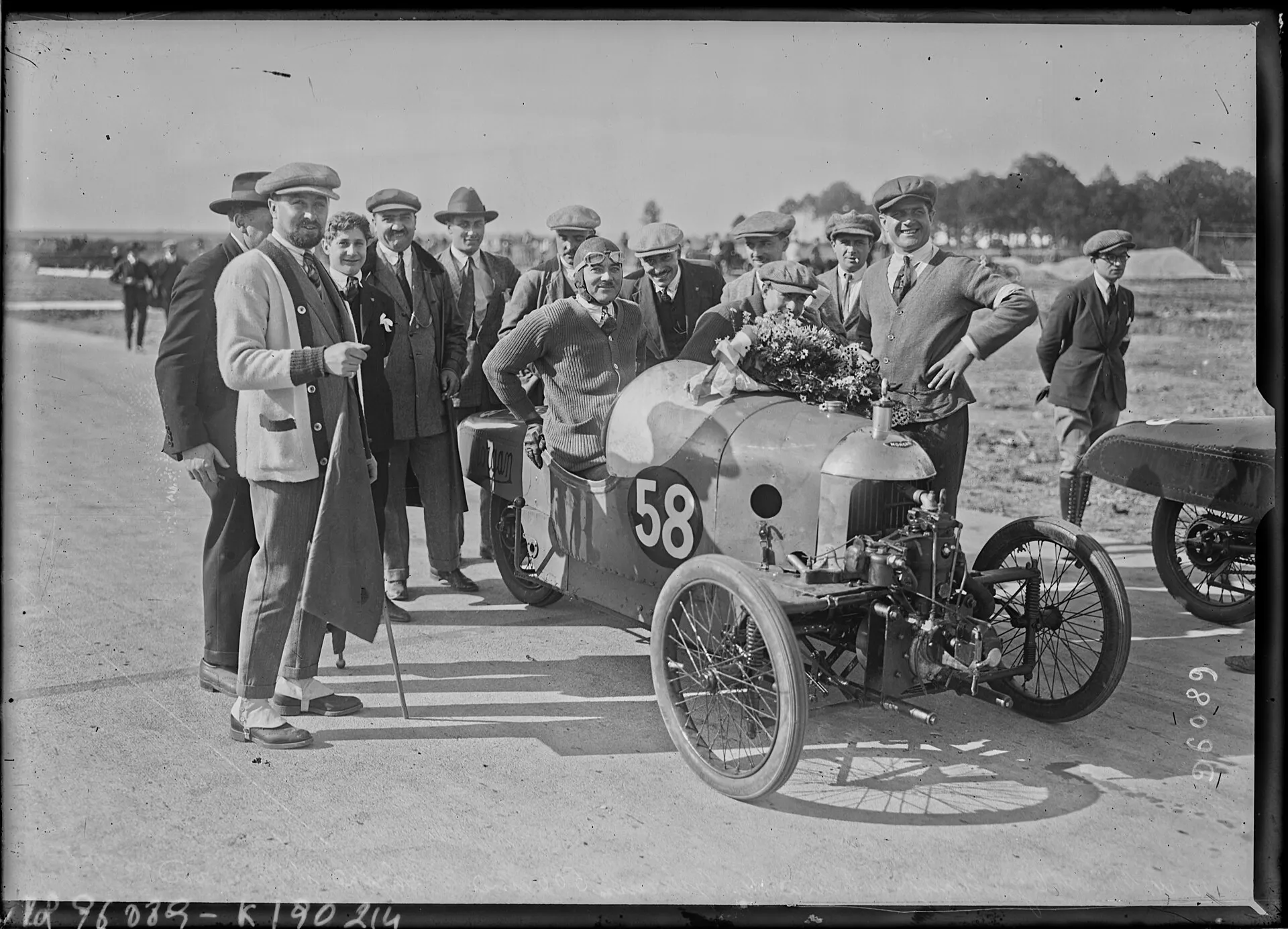 A black-and-white press photograph of a Morgan cyclecar, number 58, decorated with a garland, surrounded by men in caps at Montlhéry