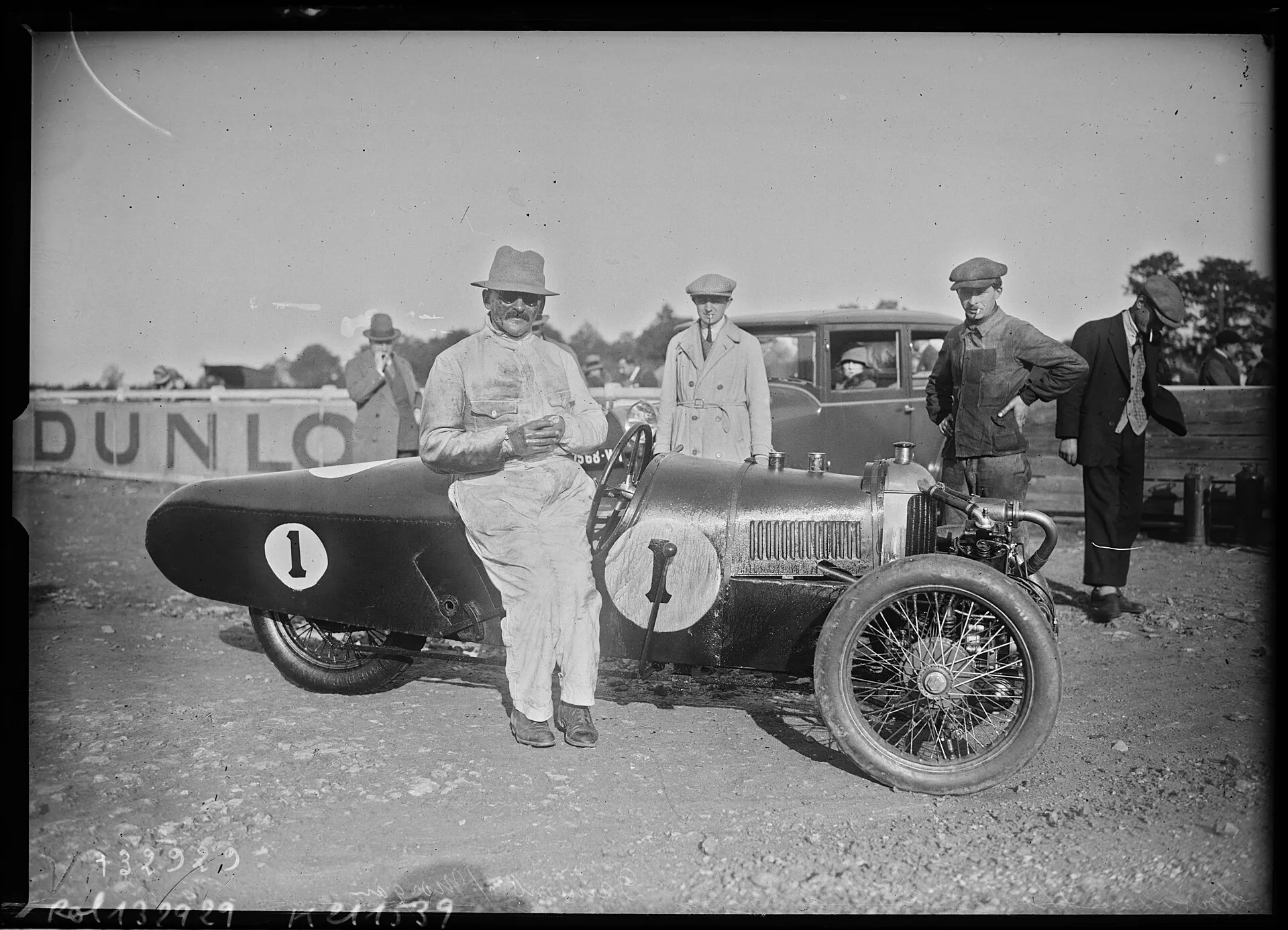 A black-and-white press photograph of a man in a light coat and hat leaning on a Morgan racing three-wheeler, number 1, at Montlhéry