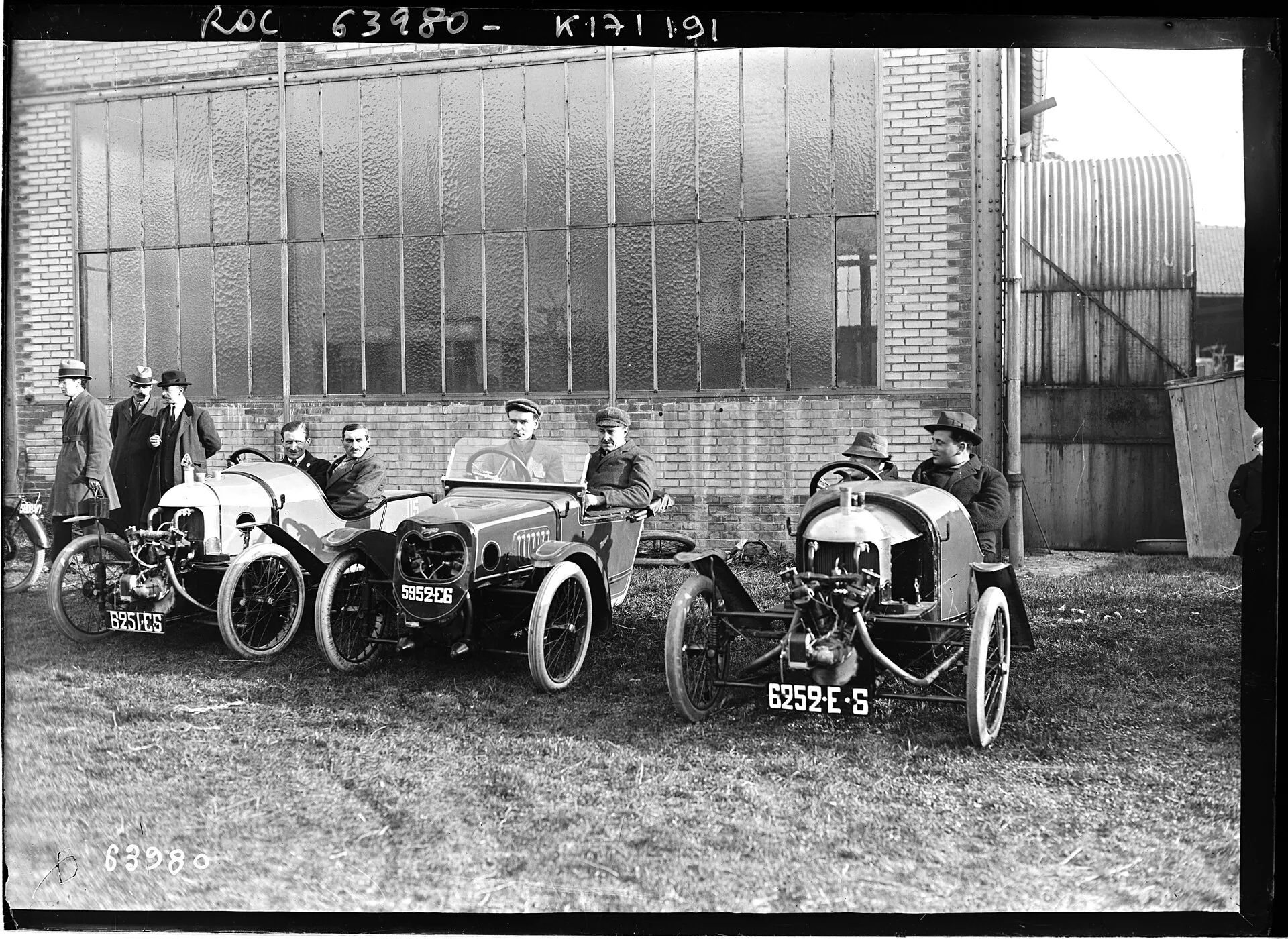A black-and-white press photograph of three Morgan cyclecars side by side in front of a hangar, with their drivers