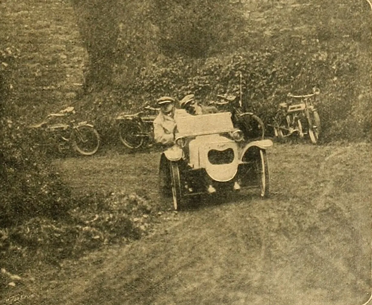 A grainy 1911 photograph of a small three-wheeled Morgan with a flat bonnet and two men in caps aboard, coming up a grassy lane between hedges, with motorcycles parked behind it