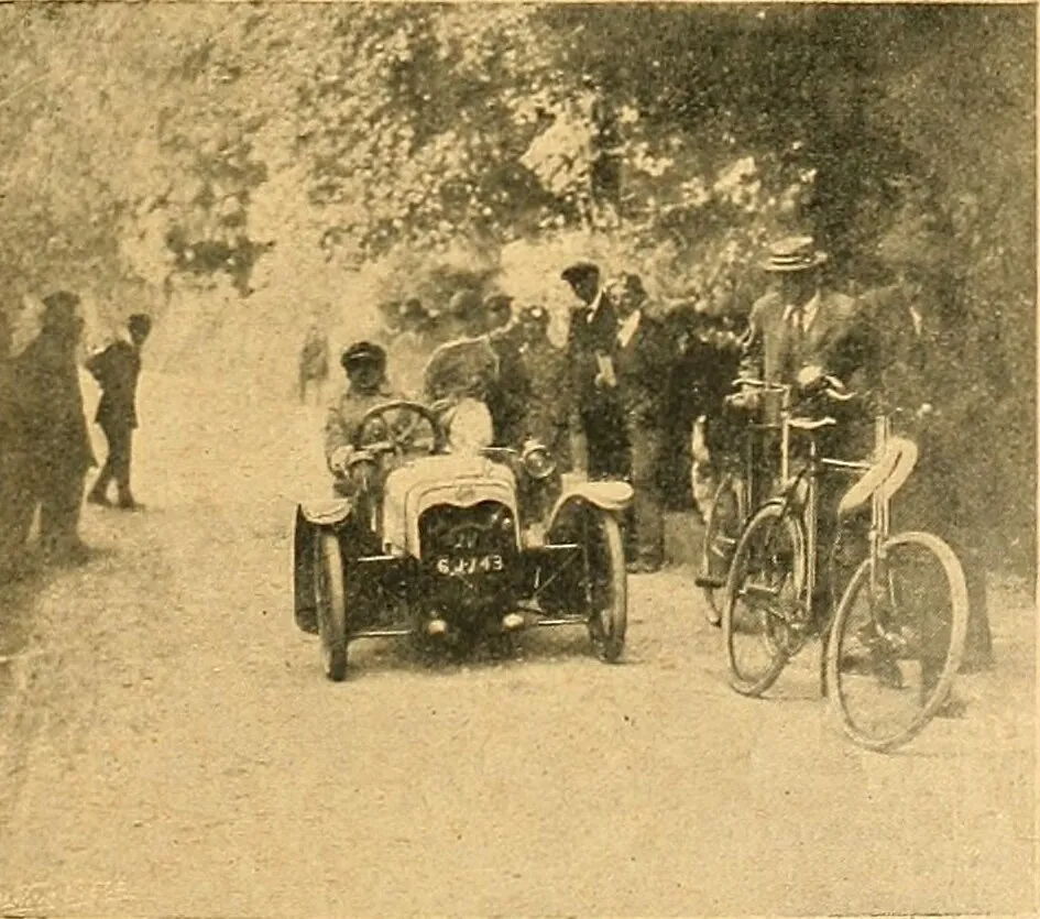 A 1911 photograph of a Morgan two-seater three-wheeler driving towards the camera on a tree-lined road, a driver in a cap at the wheel, with spectators and bicycles beside it