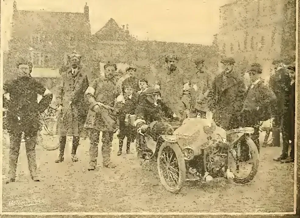A grainy 1911 photograph of a small three-wheeled Morgan, its driver in a cap leaning on the wheel, in a market square at Thirsk, with a crowd of men and boys in caps and overcoats standing round it