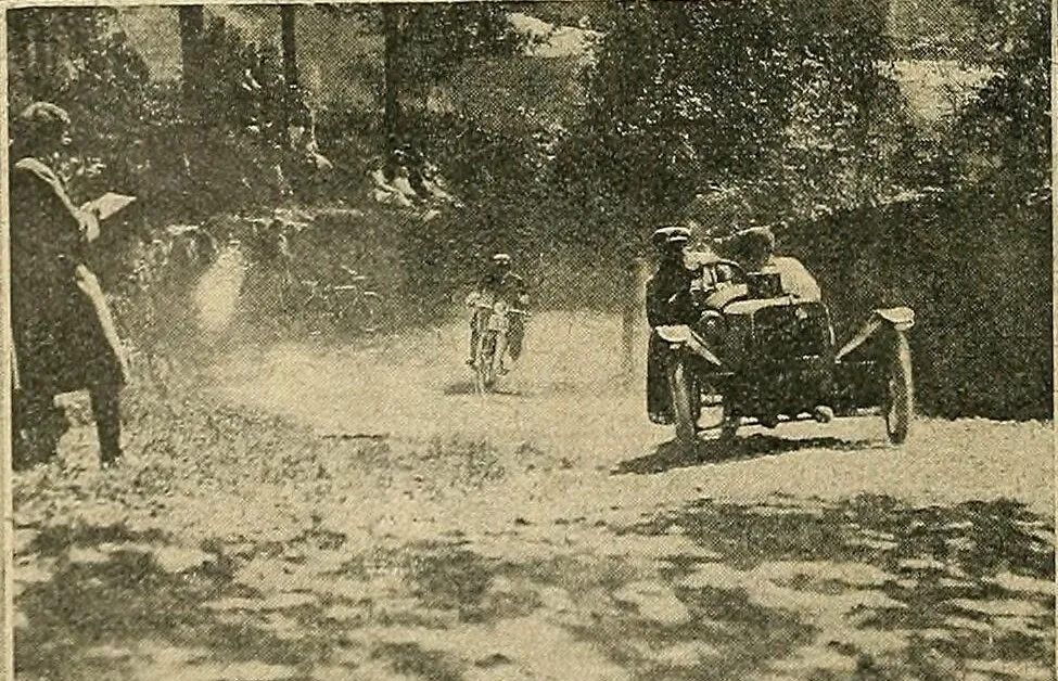 A grainy 1913 photograph of a Morgan three-wheeler with two people aboard coming up a dusty, shaded country lane, a motorcyclist behind it and an official with a notebook at the roadside
